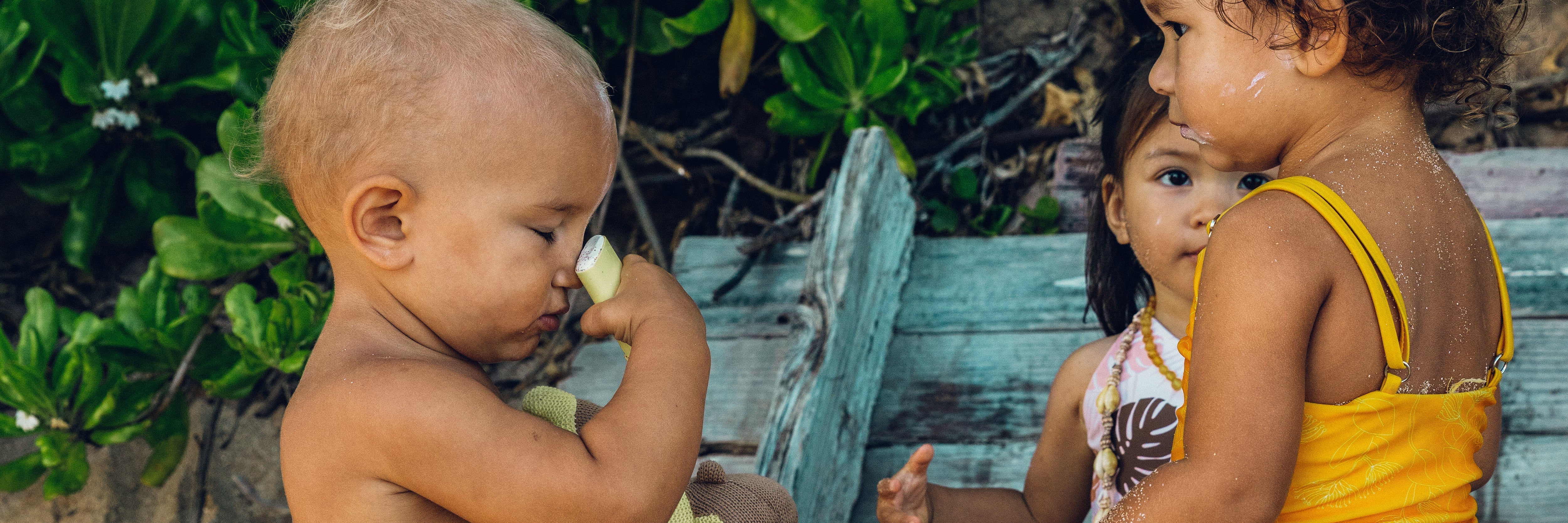 Two children playing outdoors with a wooden box and greenery in the background