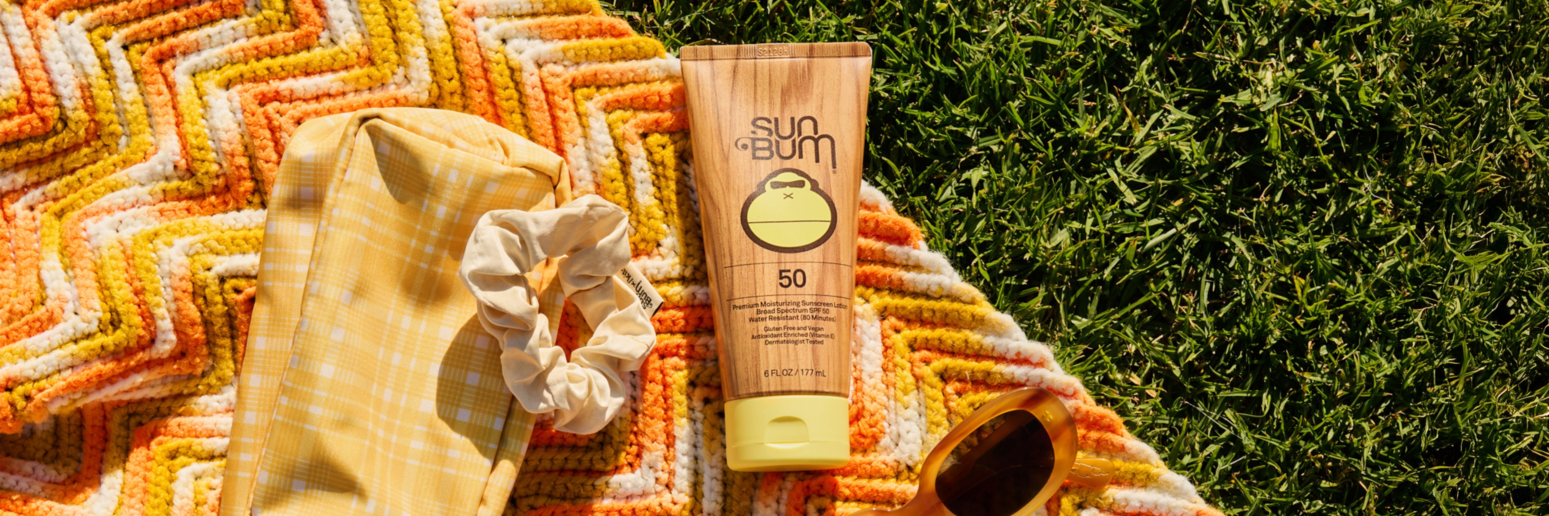 Sun Bum sunscreen tube on a colorful towel with sunglasses and a scrunchie in the grass