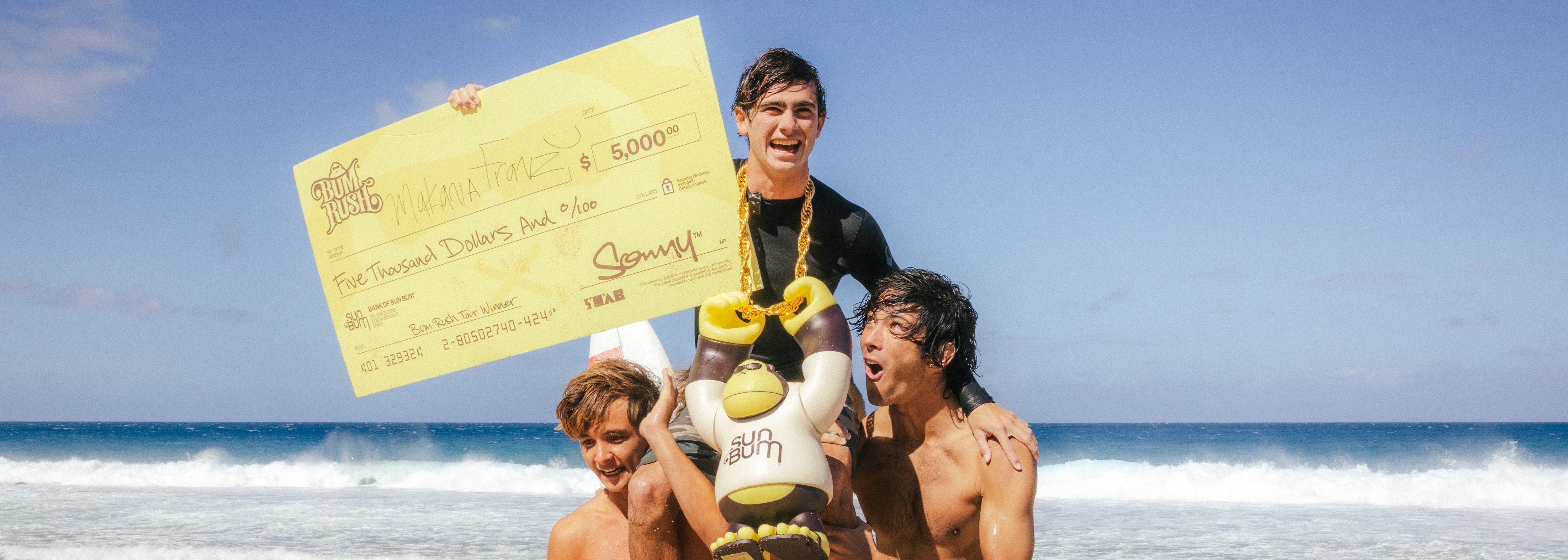 Three people on a beach with one holding a large check and another holding a toy, with ocean waves and sky in the background.