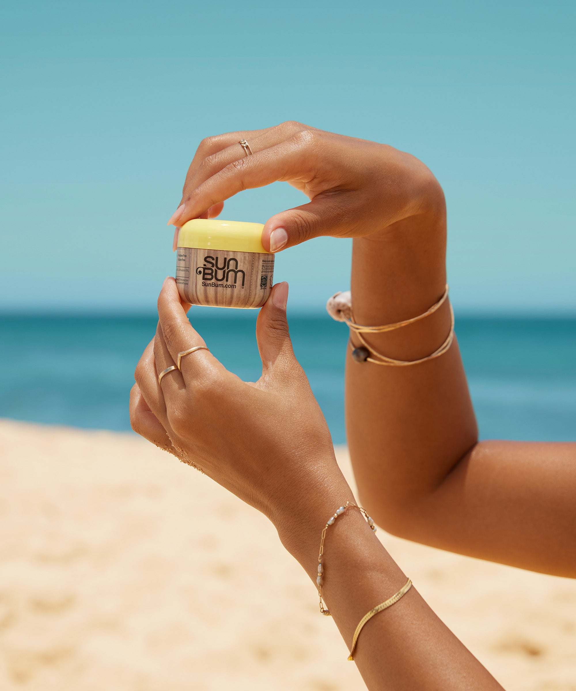 Person holding a jar of Sun Bum sunscreen on a beach with blue sky and ocean in the background.