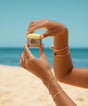 Person holding a jar of Sun Bum sunscreen on a beach with blue sky and ocean in the background.