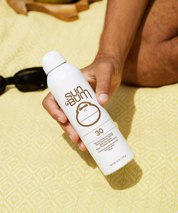 Person holding a Sun Bum sunscreen spray bottle on a yellow towel
