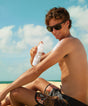Man applying sunscreen spray on a beach with a clear sky
