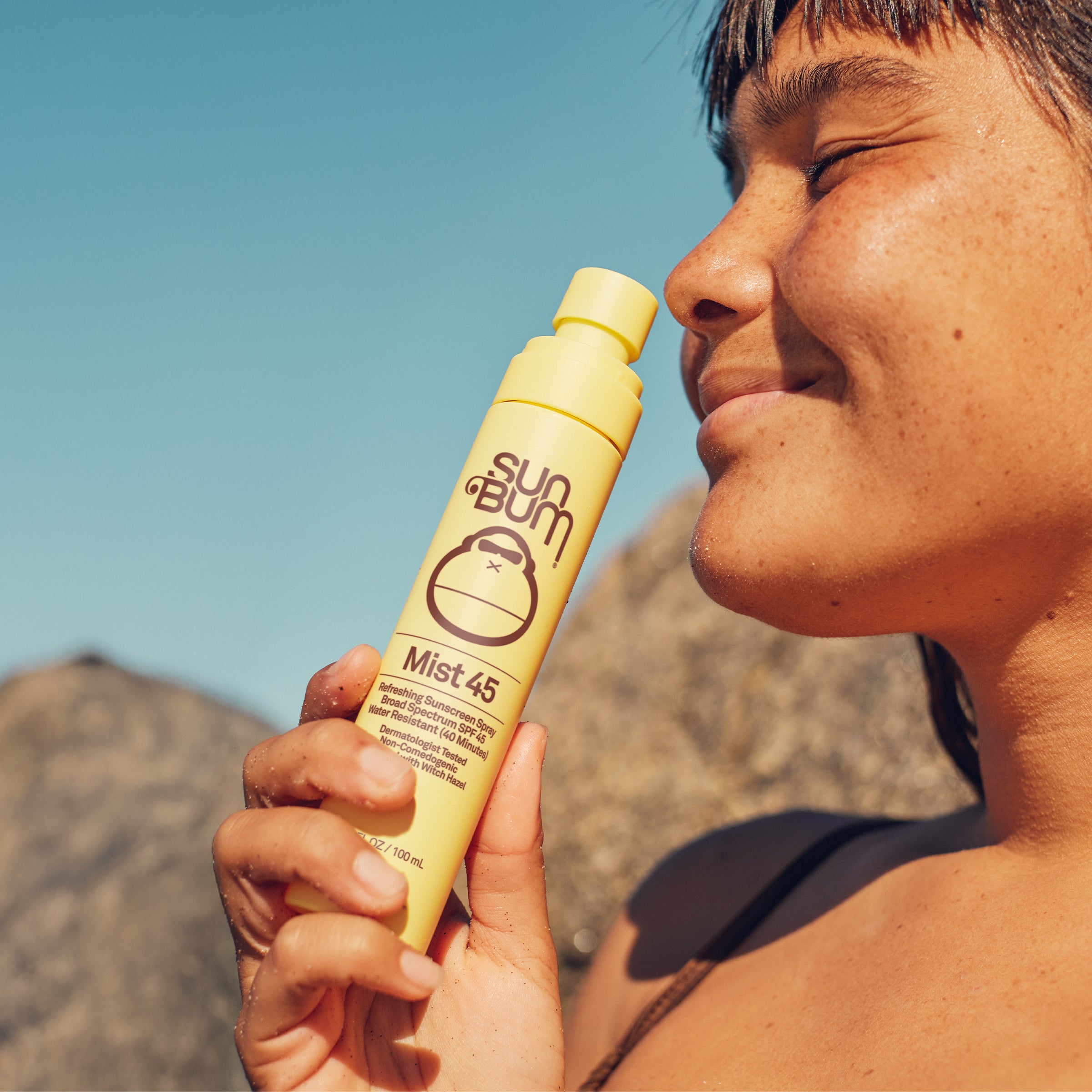 Person holding a yellow Sun Bum Mist 45 sunscreen bottle against a clear blue sky.
