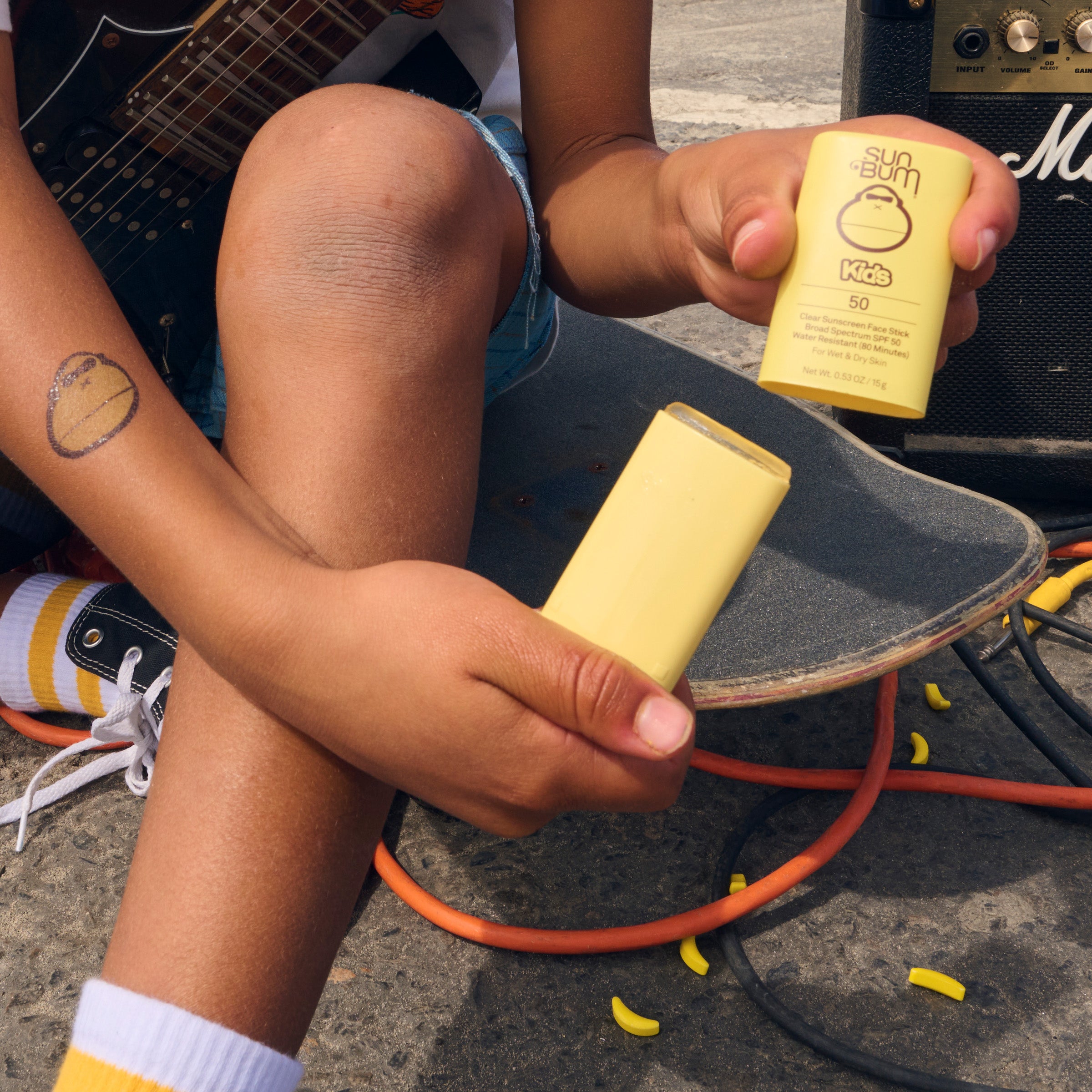 Person sitting on a skateboard holding two yellow containers, one open, with a guitar and amplifier in the background.