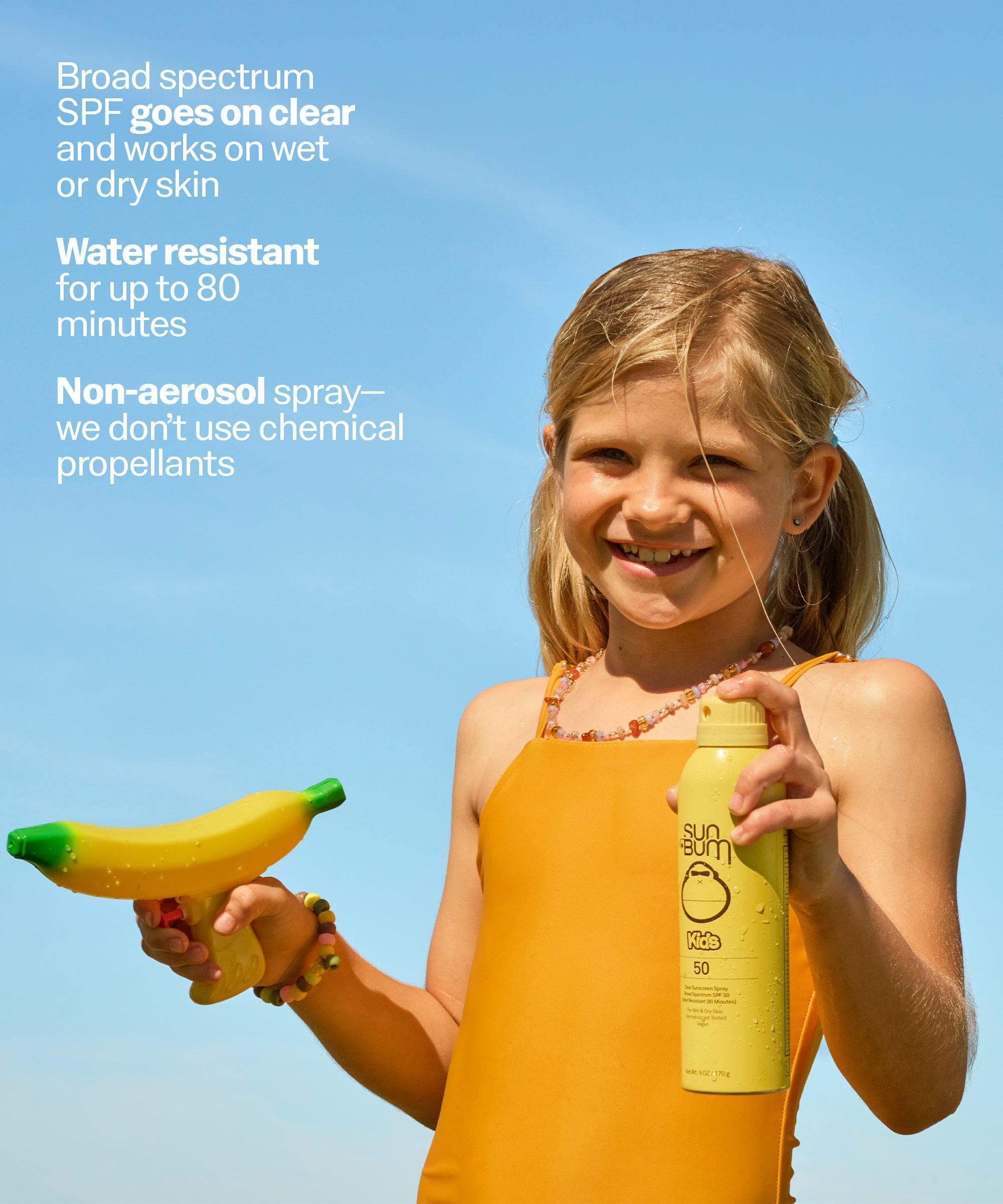 Child holding a banana and a Sun Bum sunscreen spray bottle against a clear blue sky.