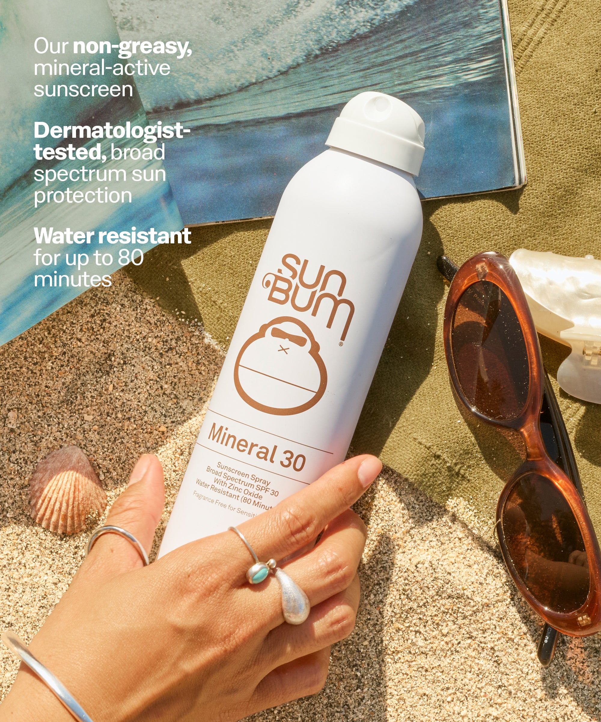 Sun Bum Mineral 30 sunscreen spray held by a hand on sand with sunglasses and shells in the background.