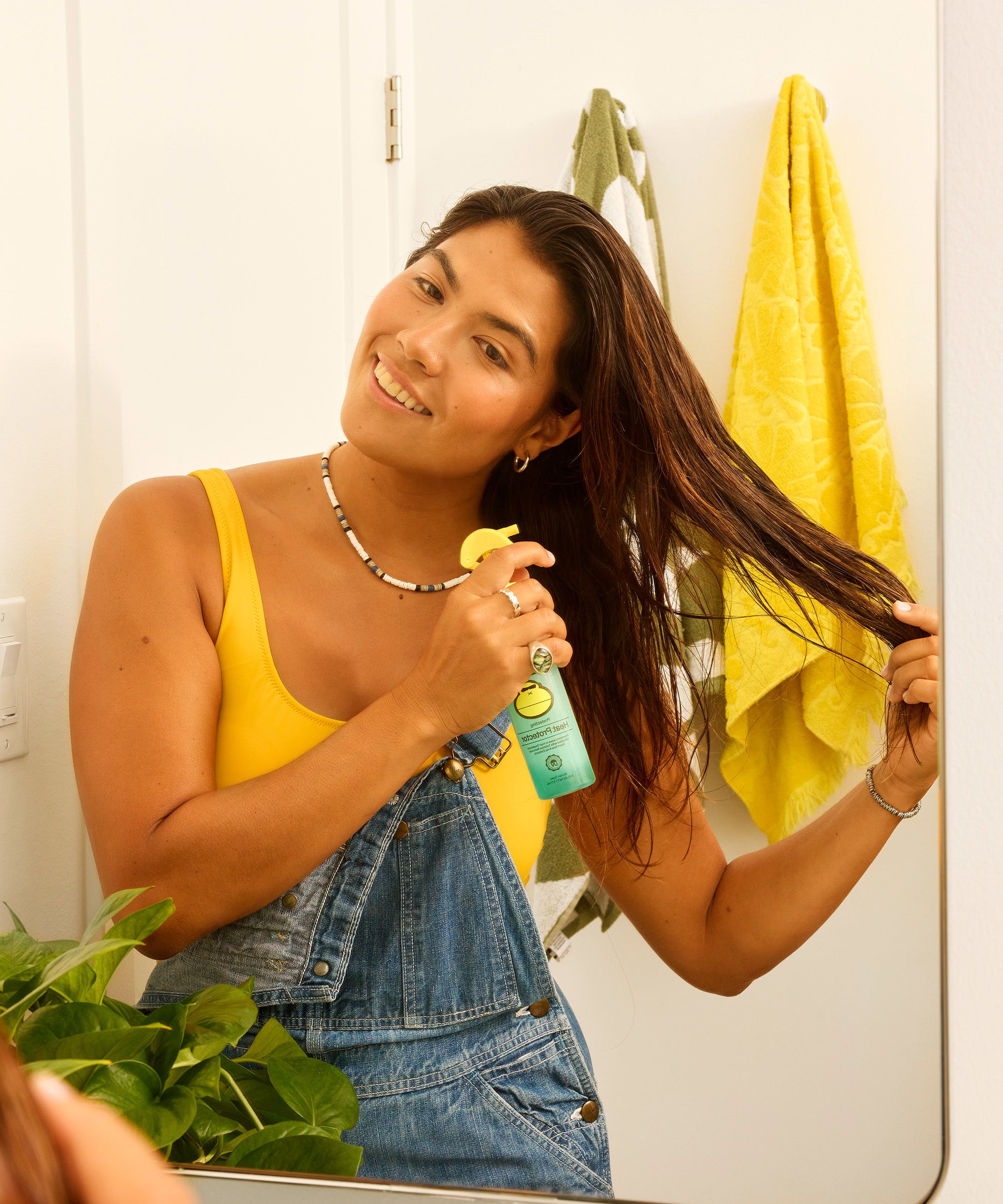 Woman in a yellow tank top and denim shorts applying hair product in front of a mirror.
