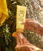 Hand holding a tube of Sun Bum Hair Mask with water droplets in the background
