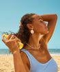 Woman applying sunscreen on a beach with a clear blue sky and ocean in the background.