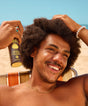 Man with curly hair holding a can of Sun Bum sunscreen spray against a clear blue sky.