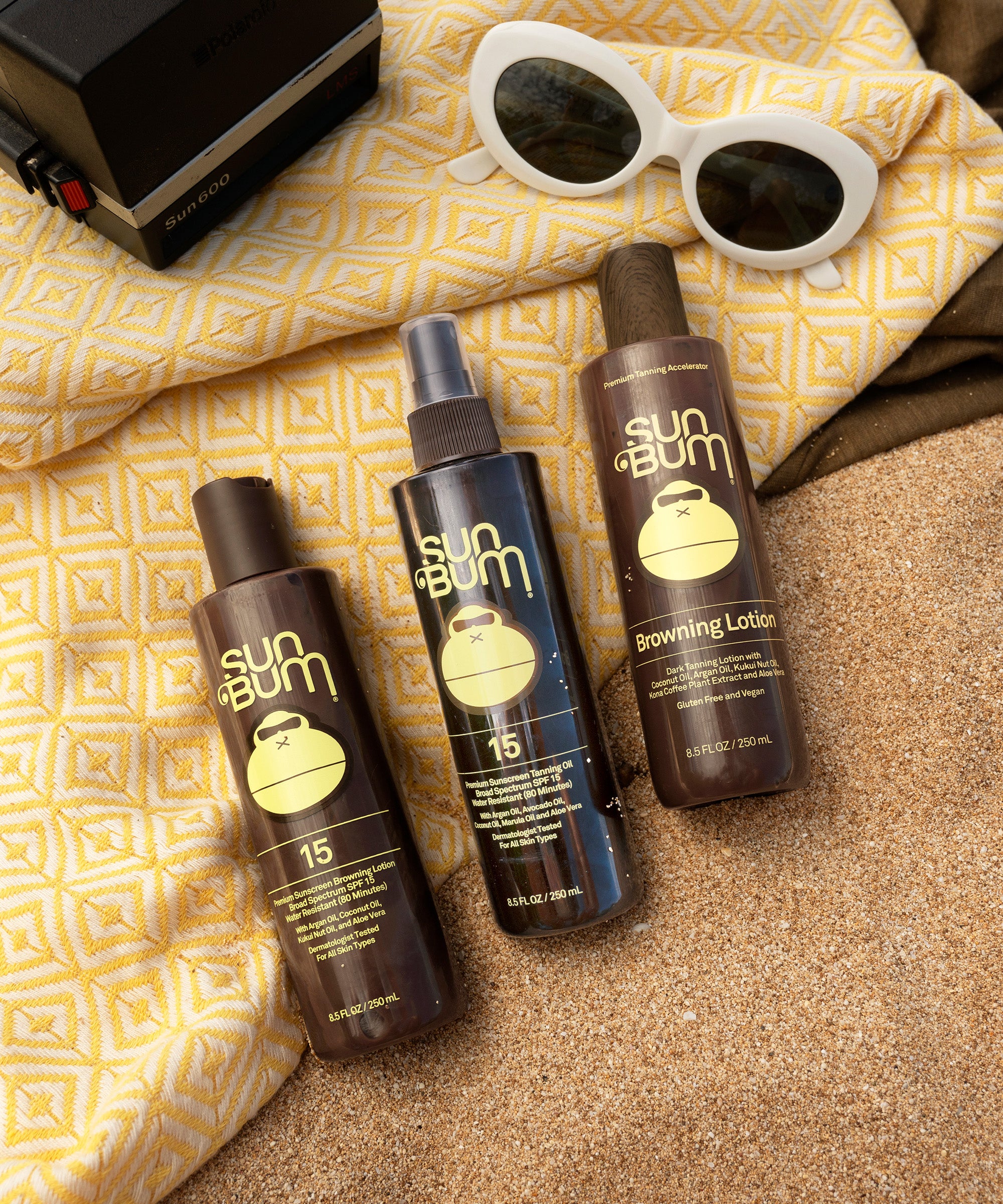 Sun Bum sunscreen products on sand with a yellow towel and sunglasses in the background