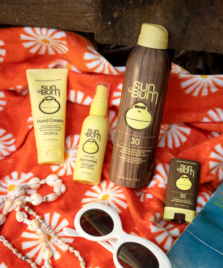 Sun Bum sunscreen products on a red floral towel with sunglasses and a necklace.
