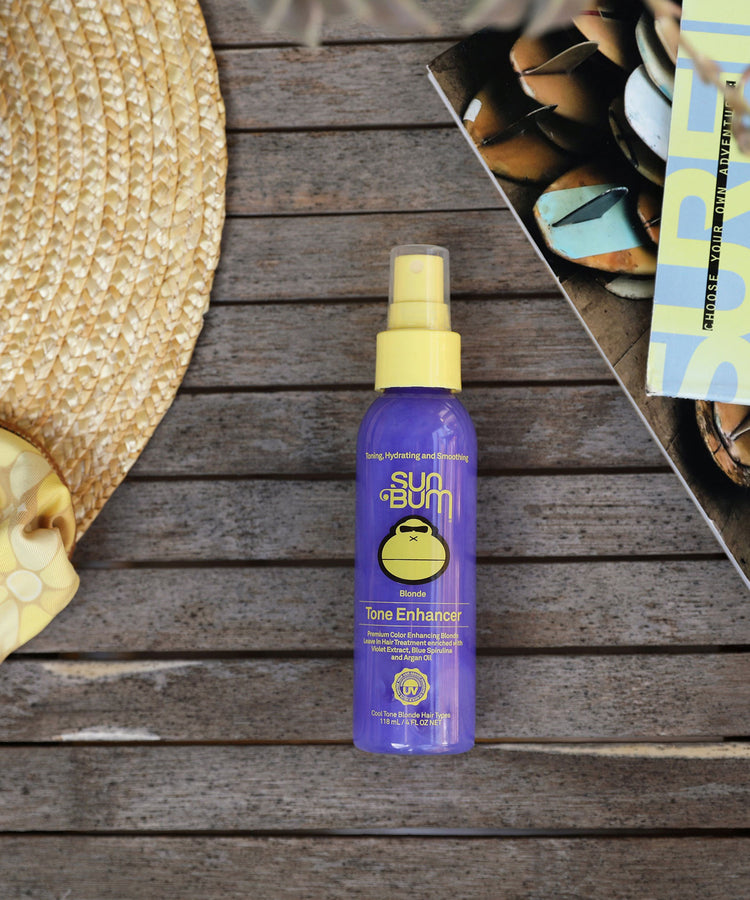 Sun Bum Tone Enhancer spray bottle on a wooden surface with a hat and towel in the background