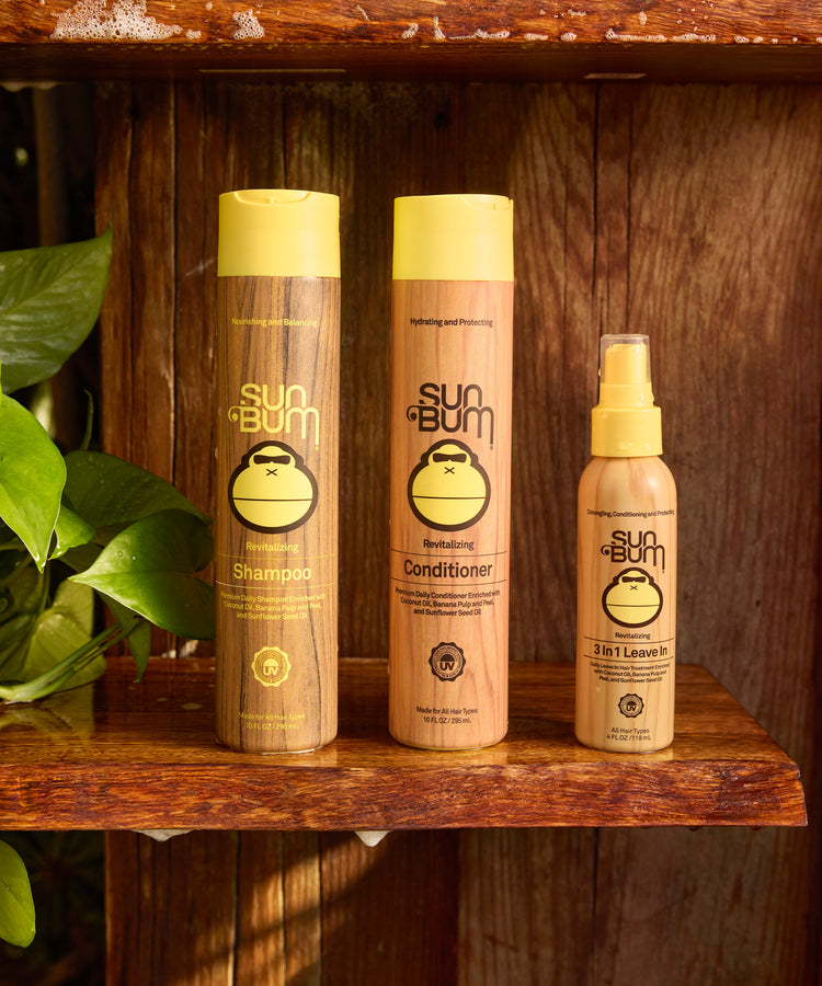 Sun Bum shampoo, conditioner, and spray bottle on a wooden shelf with a plant in the background.