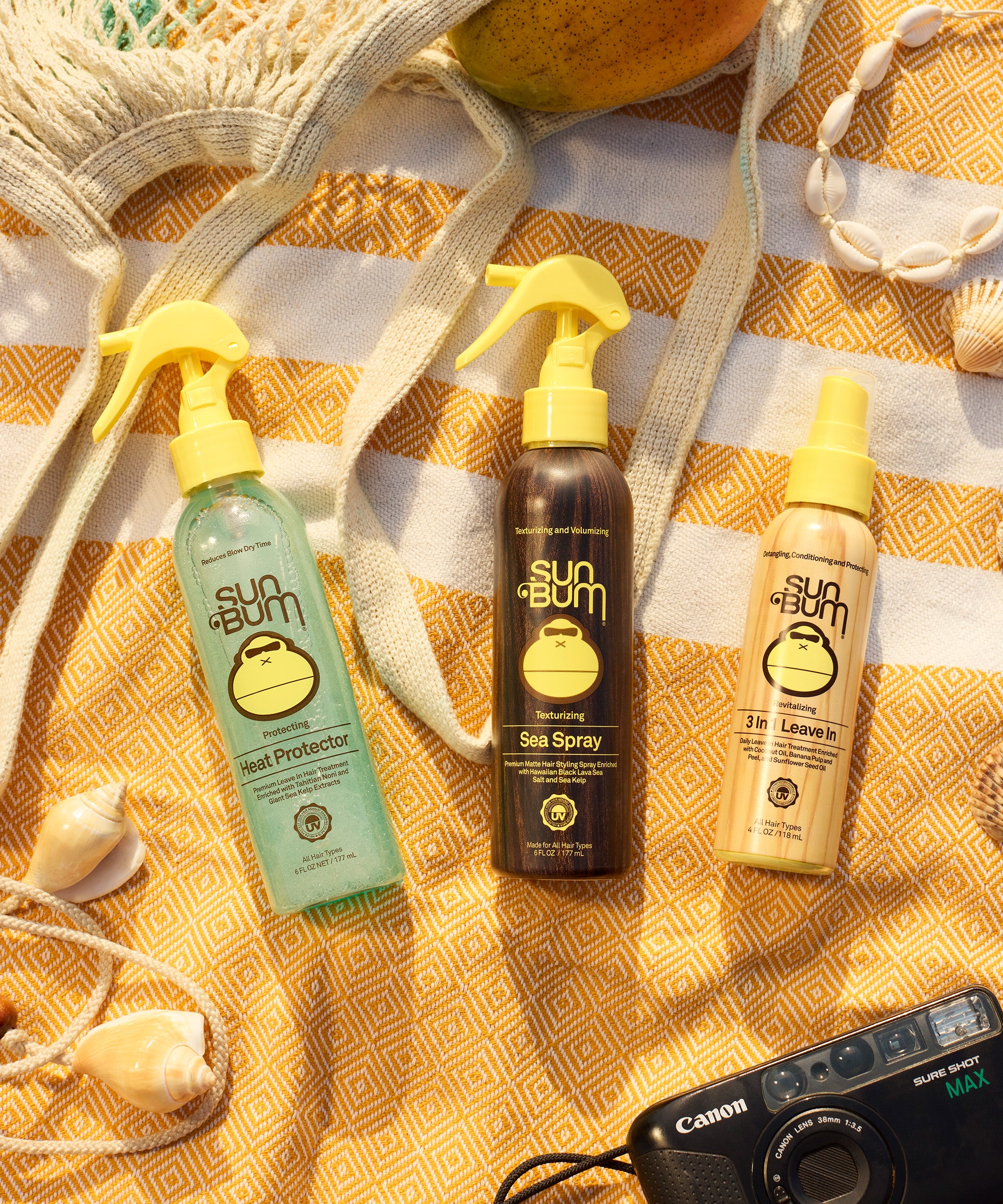 Three Sun Bum sunscreen products on a yellow patterned towel with a camera and seashells.