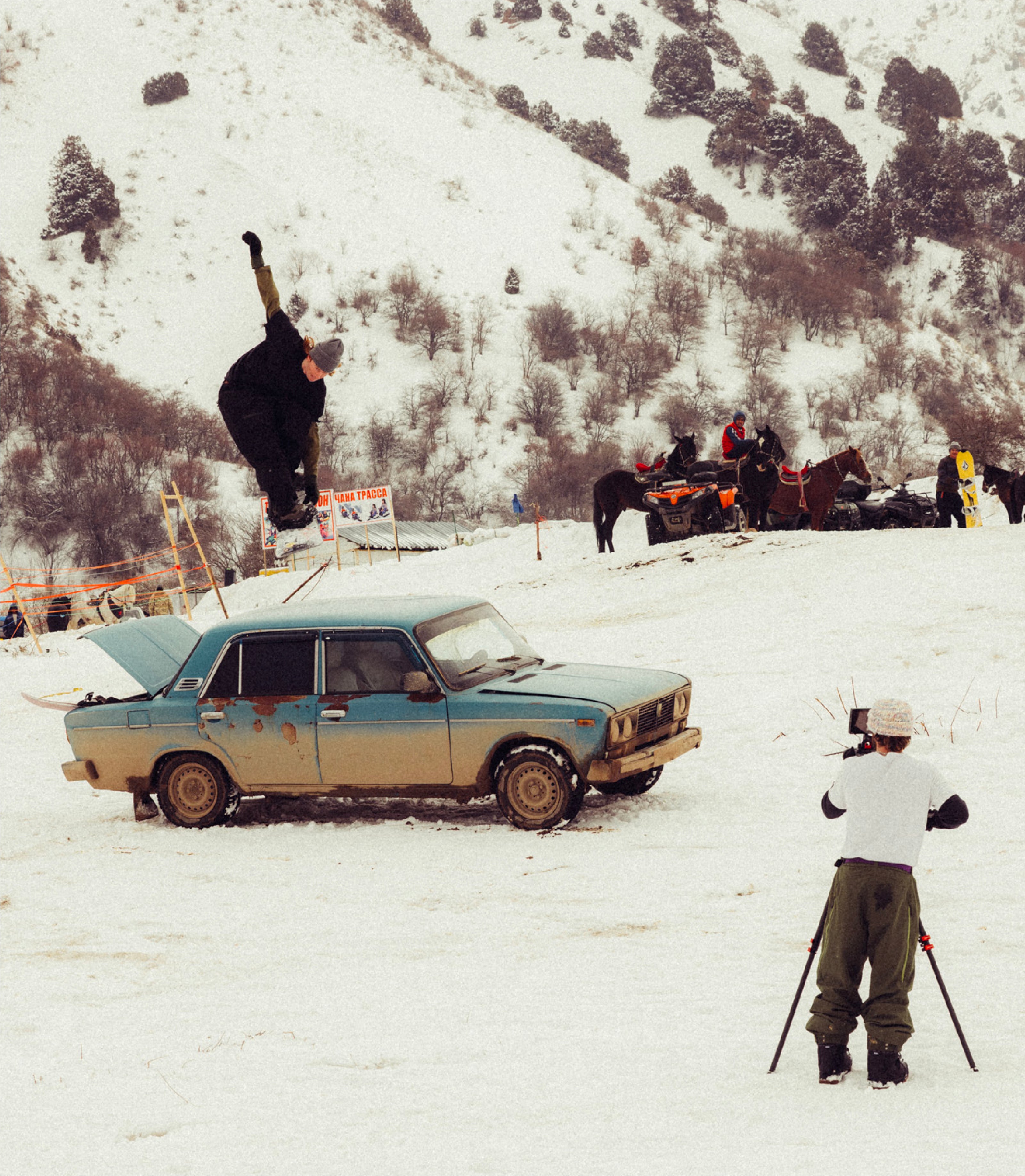 Person performing a snowboard trick over an old car in a snowy landscape with onlookers and photographers.