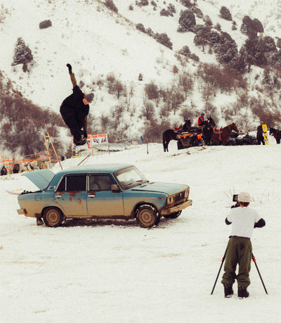 Person performing a snowboard trick over an old car in a snowy landscape with onlookers and photographers.