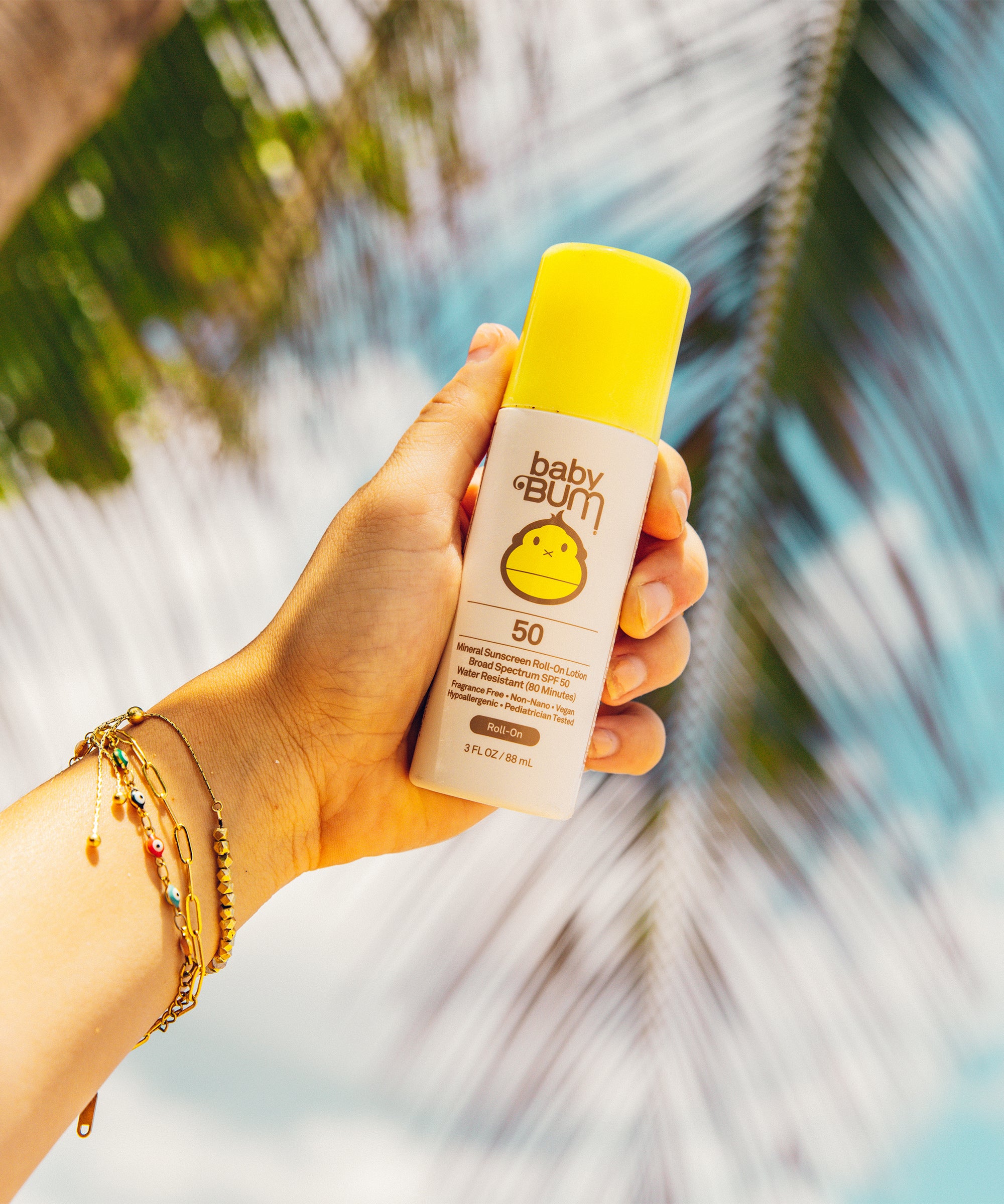 Hand holding a bottle of Baby Bum sunscreen with palm leaves in the background