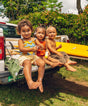 Three children sitting on a truck bed with a yellow kayak, surrounded by greenery.
