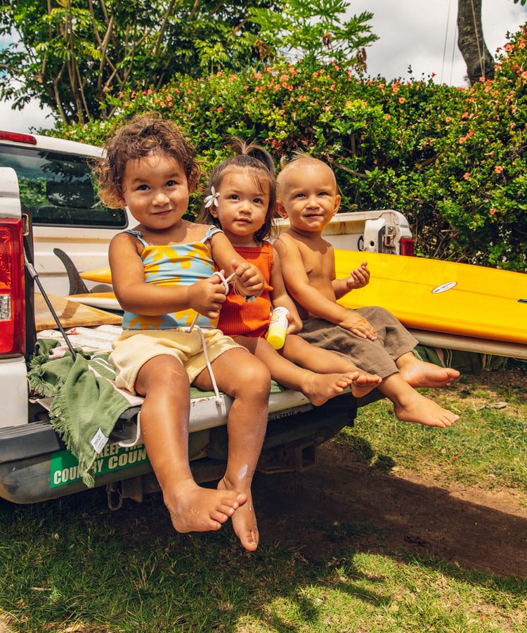 Three children sitting on a truck bed with a yellow kayak, surrounded by greenery.