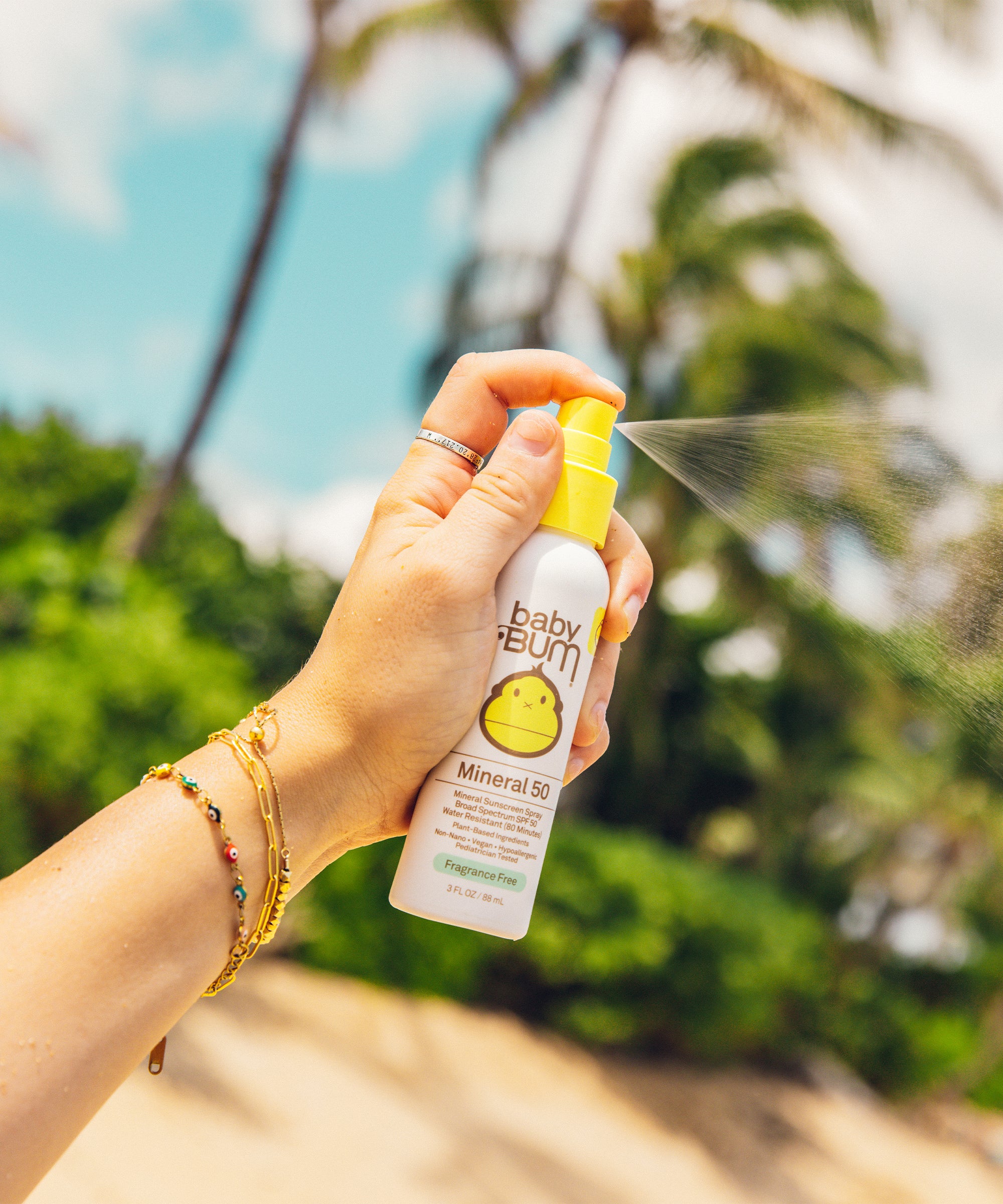 Person holding a Baby Bum sunscreen spray bottle with palm trees in the background