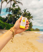 Hand holding a sunscreen bottle with palm trees and beach in the background