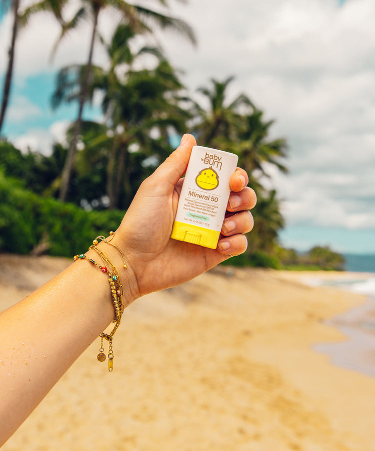 Hand holding a sunscreen bottle with palm trees and beach in the background