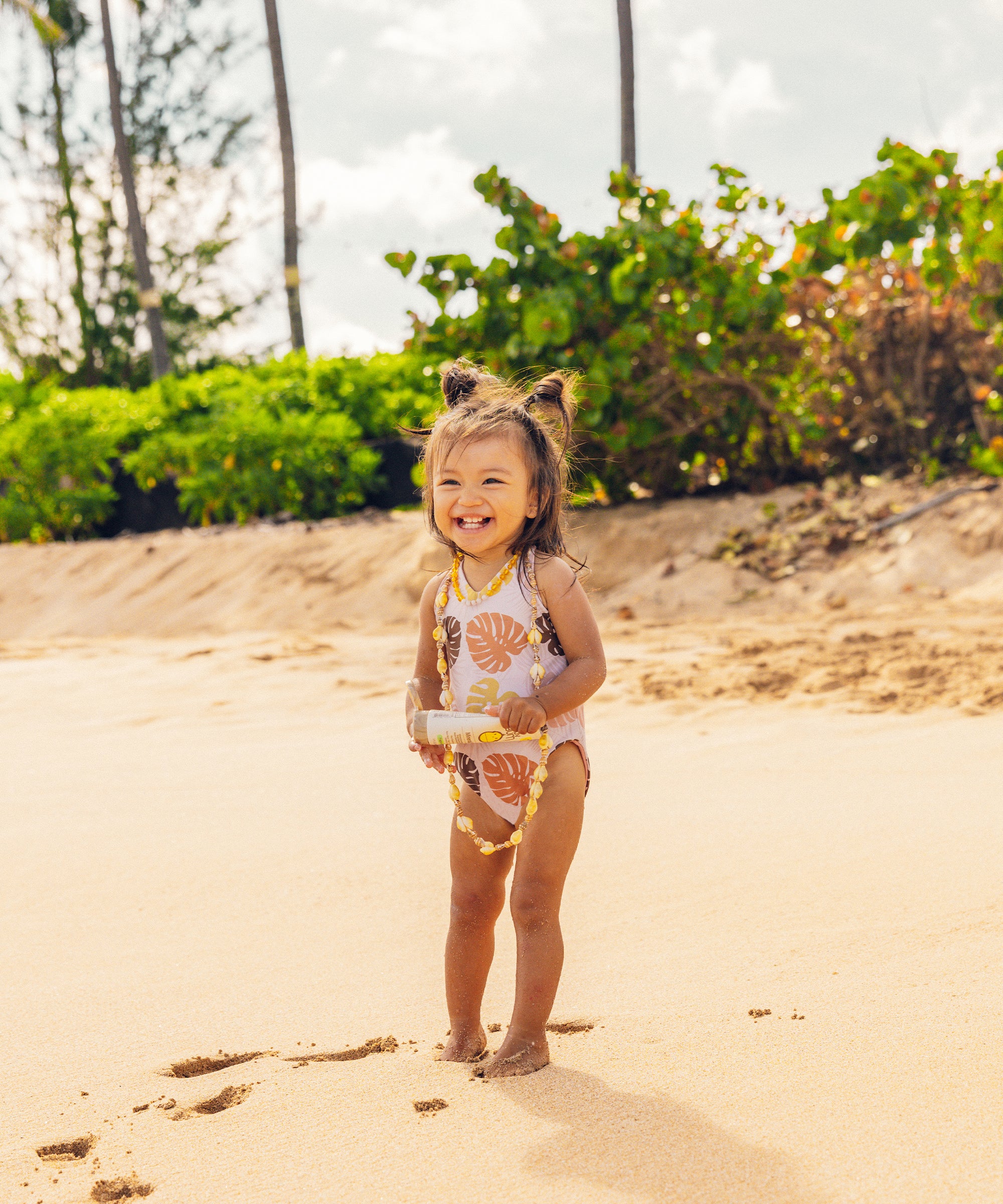 Child in a floral swimsuit standing on a sandy beach with greenery in the background