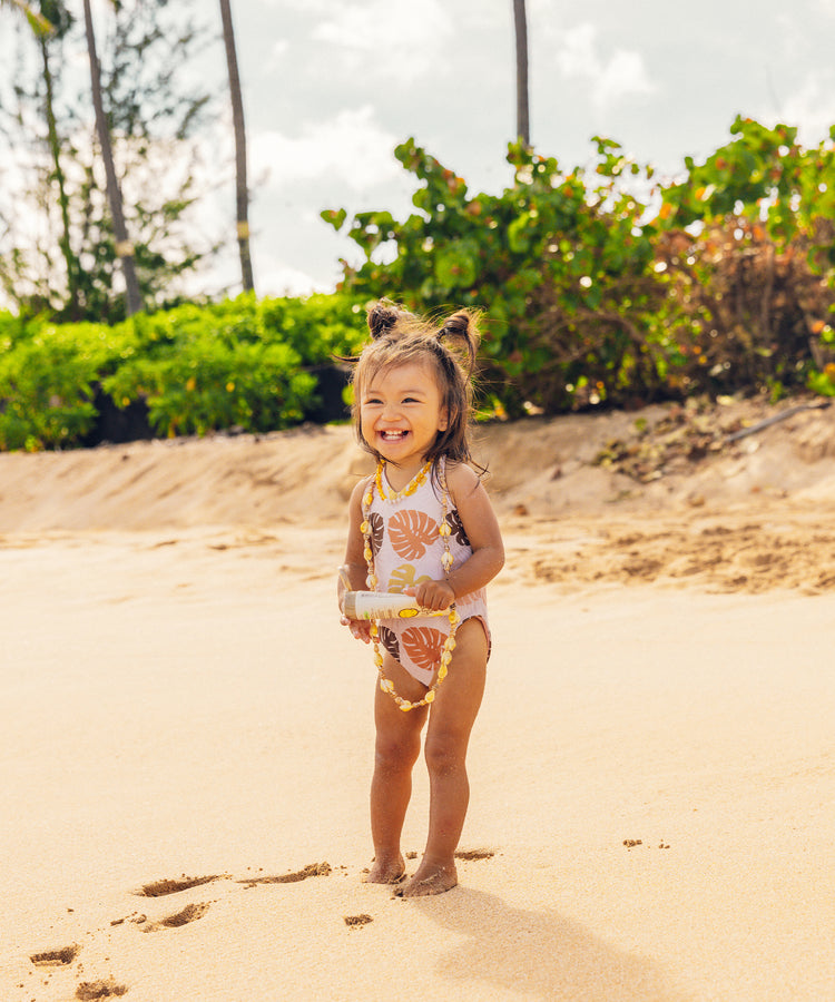 Child in a floral swimsuit standing on a sandy beach with greenery in the background