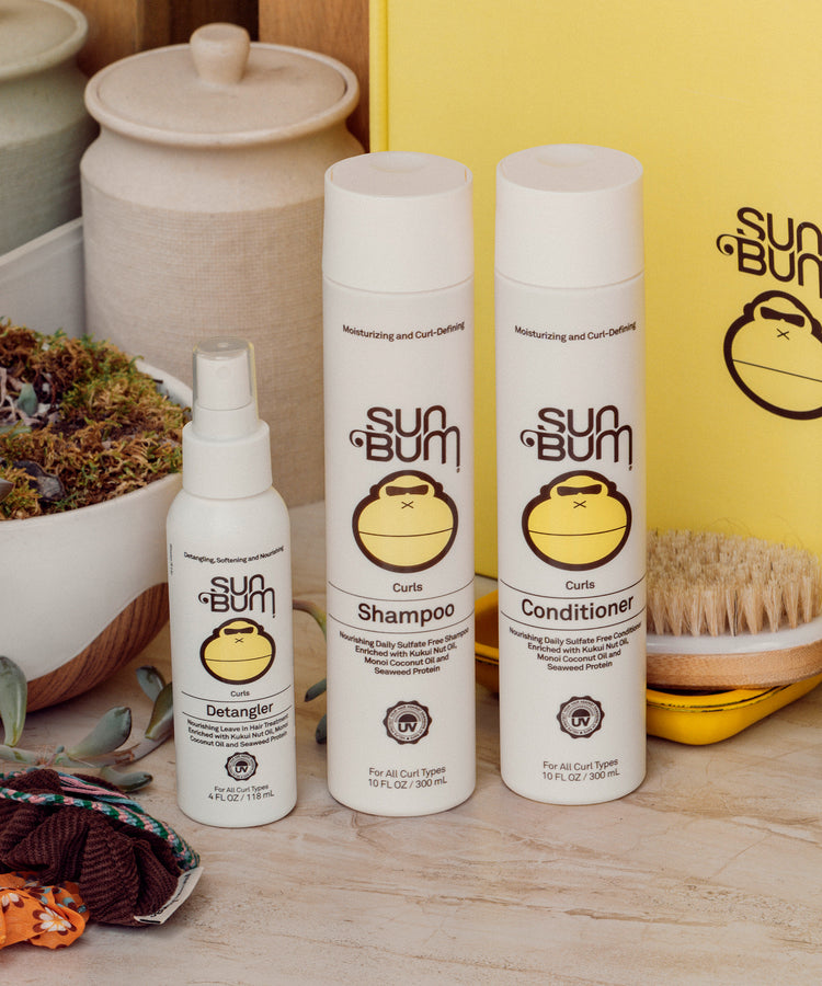 Sun Bum hair care products including shampoo, conditioner, and detangler on a wooden surface with a yellow box in the background.