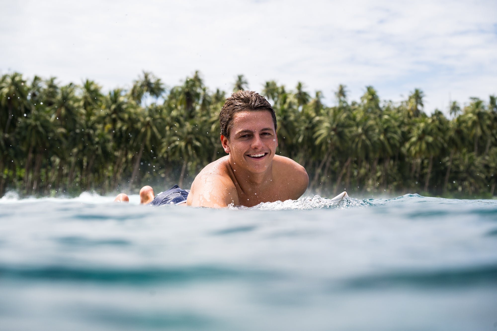 Man lying on a surfboard in the water with palm trees in the background