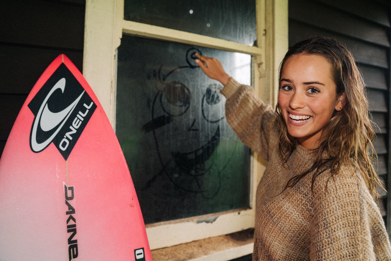 Woman standing next to a pink surfboard with O'Neill logo, pointing at a window.