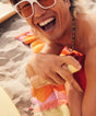 Person on a beach holding a shell, wearing sunglasses and a colorful towel.