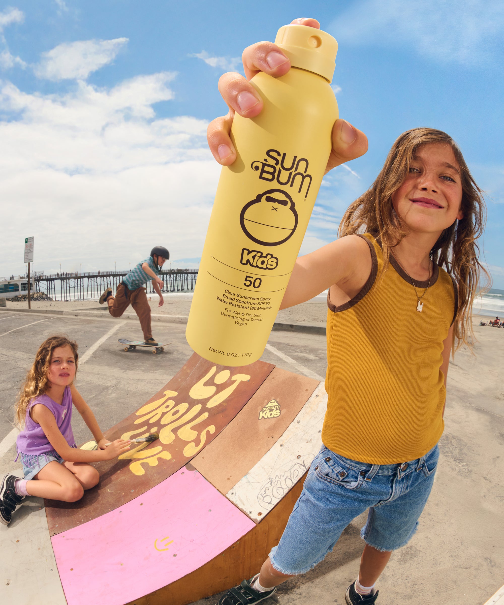 Child holding a Sun Bum Kids sunscreen bottle on a beach with another child in the background.