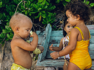 Two children playing outdoors with a wooden box and greenery in the background