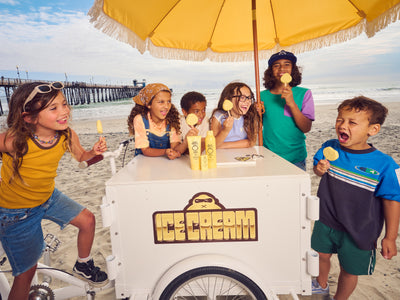 Children selling ice cream from a cart on a beach with a yellow umbrella.