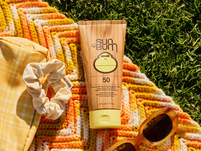 Sun Bum sunscreen tube on a colorful towel with sunglasses and a scrunchie in the grass