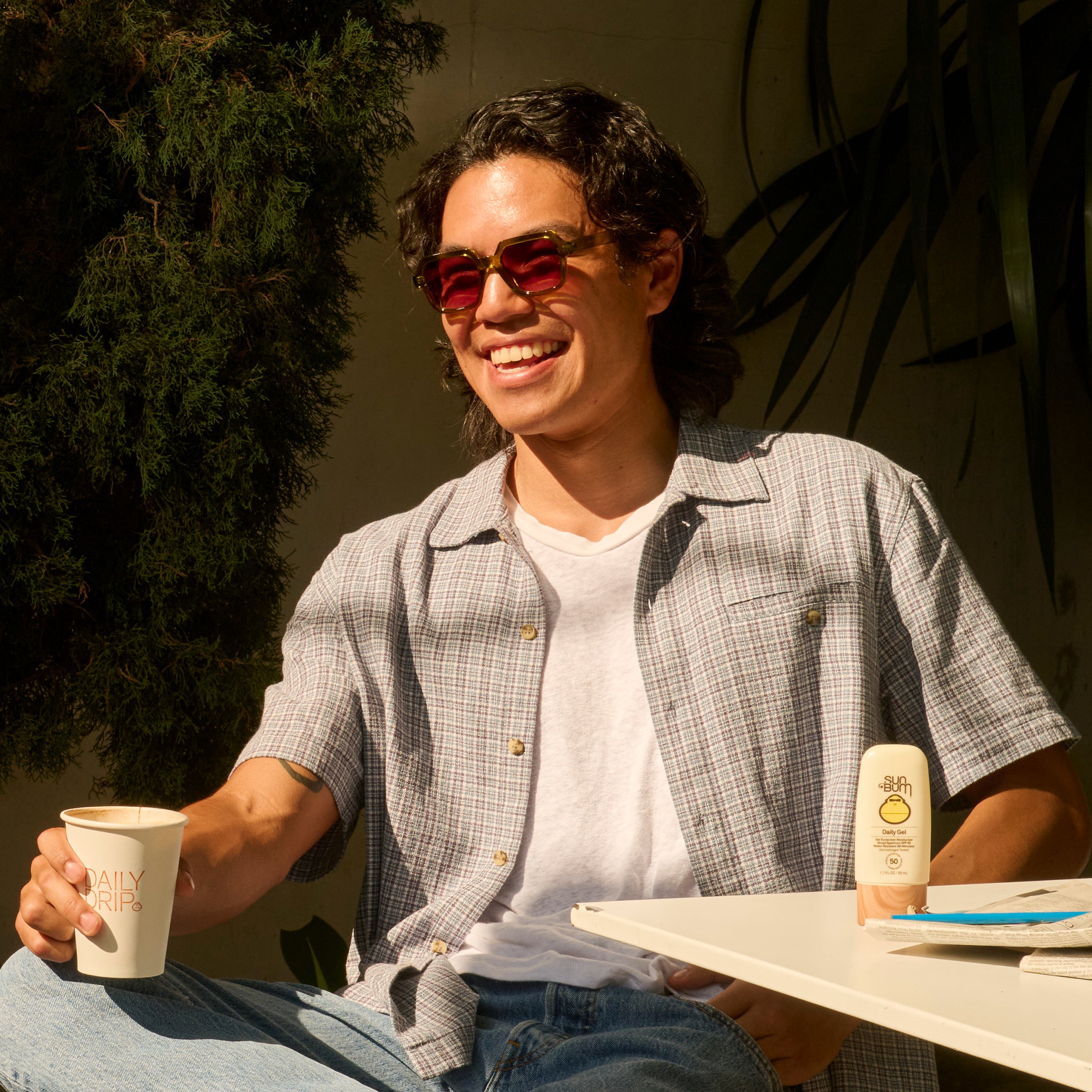 Man sitting outdoors holding a coffee cup, wearing sunglasses and a plaid shirt.