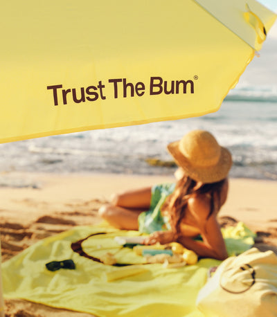 Person sitting on a beach with a yellow 'Trust The Bum' umbrella and towel.