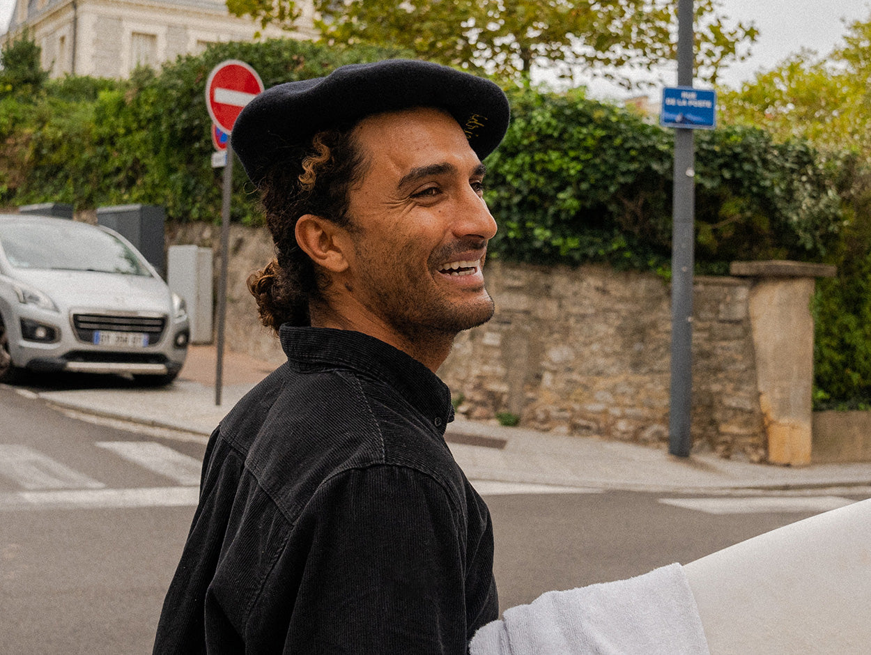 Man in black cap and dark shirt standing on a street with cars and greenery in the background