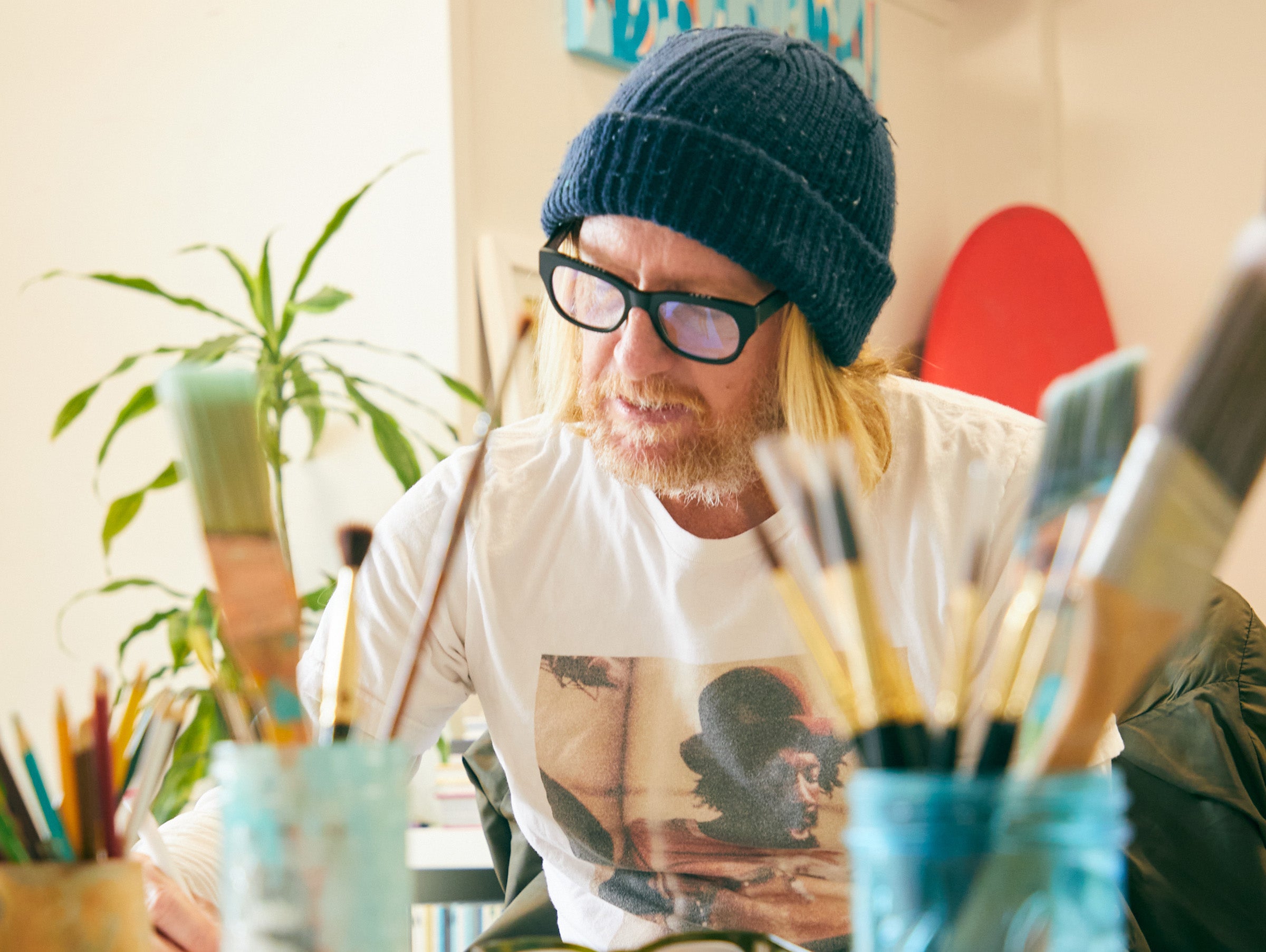 Man with glasses and a beanie surrounded by paintbrushes and art supplies