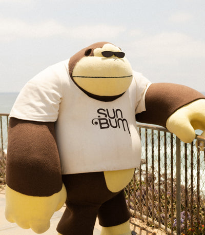 Stuffed monkey toy wearing a 'Sun Bum' shirt outdoors.