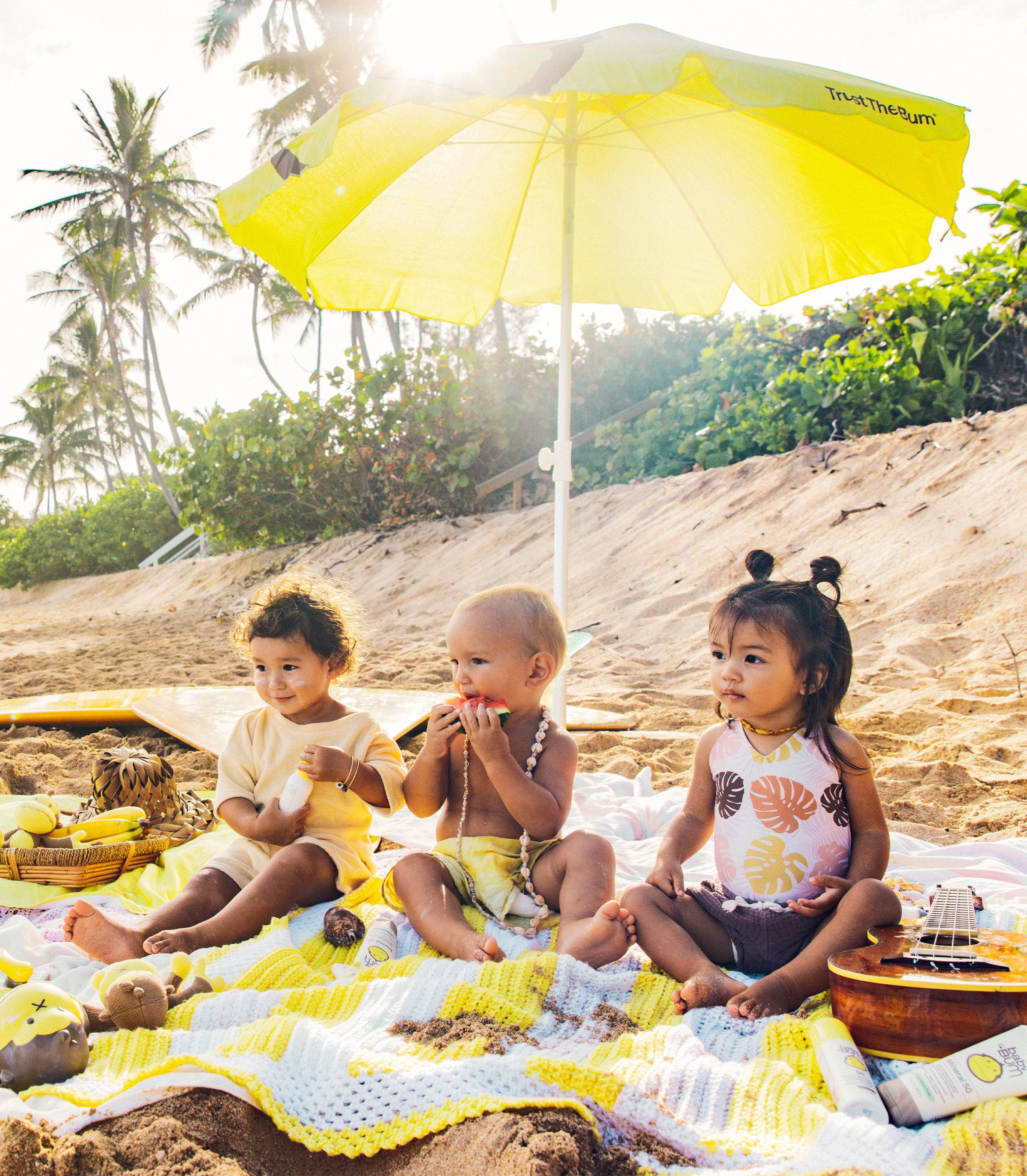 Three children sitting on a blanket under a yellow umbrella on a beach.