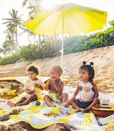 Three children sitting on a blanket under a yellow umbrella on a beach.