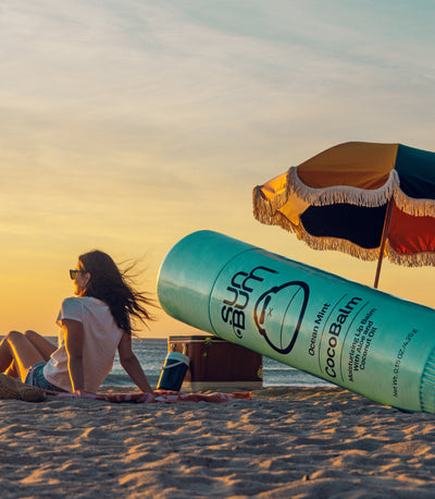Woman sitting on a beach with a colorful umbrella, and a large bottle of Suu Balm in the foreground.