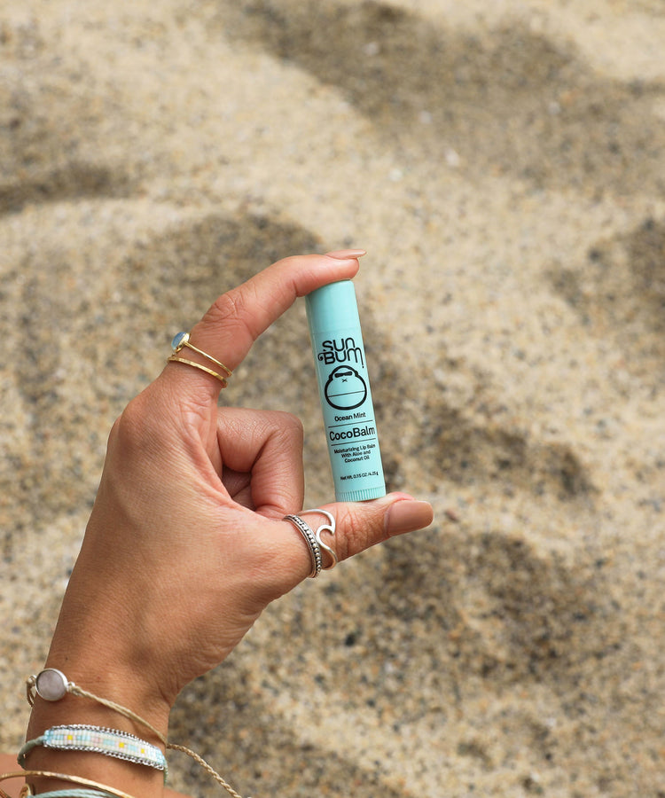 Hand holding a blue tube of sunscreen balm against a sandy background
