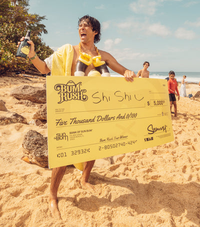 Person on a beach holding a large check with 'Bum Rush' branding