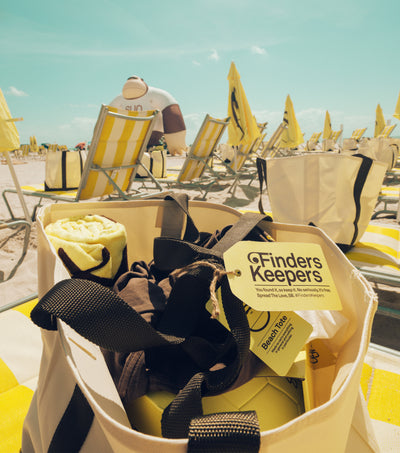 Beach bag with accessories on a sandy beach with yellow and white chairs.