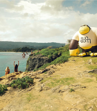 Inflatable sunbrella character on a cliff overlooking a scenic landscape with people jumping into the water.